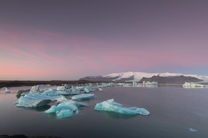 This timelapse video captures the transition from twilight to early morning over a glacial lagoon filled with luminous blue icebergs as the sky shifts from a deep indigo to a soft pastel pink and eventually a bright clear daylight the calm water reflec
