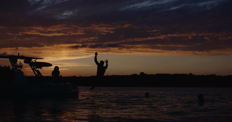 The silhouette of a man jumps from the back of a boat into a lake as a woman watches during a vibrant sunset with orange and dark purple clouds in the sky