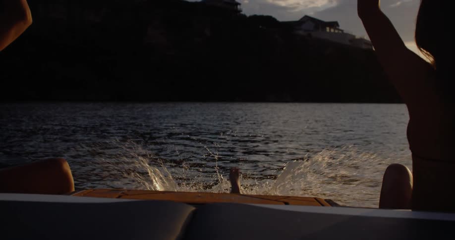 A man in swimming trunks dives from the back of a boat into a lake as two women in bikinis watch from the deck during a dark sunset with orange clouds on the horizon