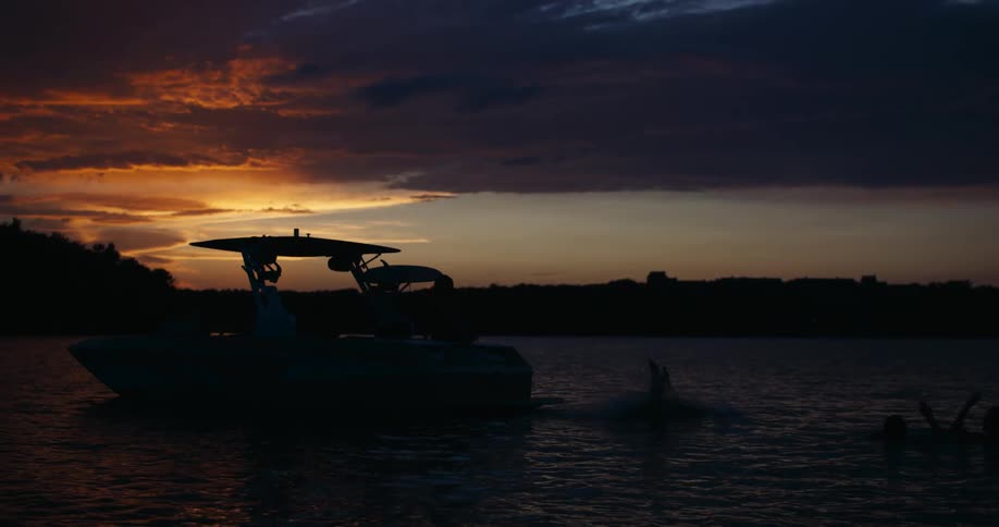 This silhouette shot a person dives headfirst off the back of a boat into a calm lake the scene is set against a dramatic sunset with orange and dark purple clouds stretching across the horizon