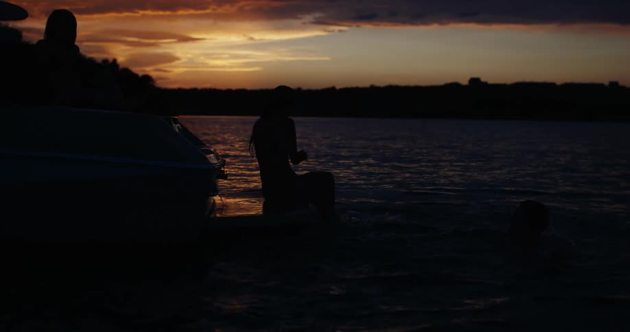 A person in swim trunks dives into the water from a boat while two women sit on the deck and watch during a dark sunset with orange clouds on the horizon