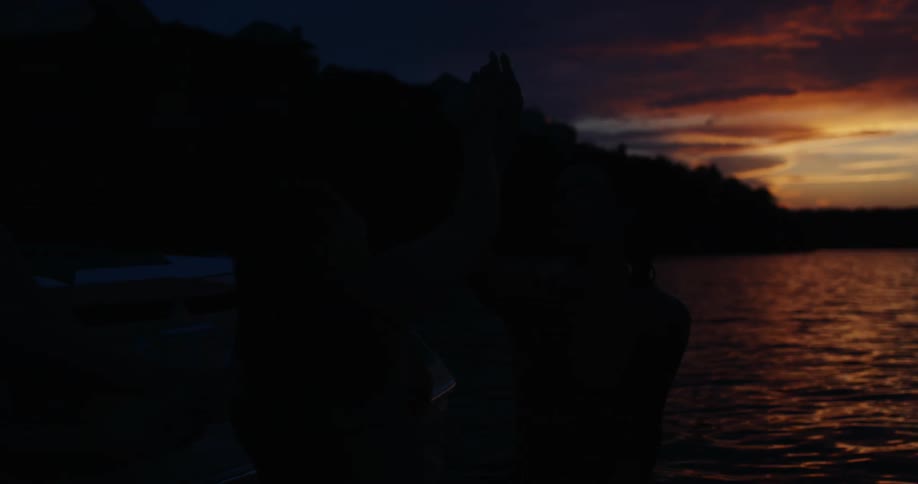 A view from the deck shows the silhouettes of two women sitting as they raise their arms and high five during a dark sunset with orange clouds on the horizon