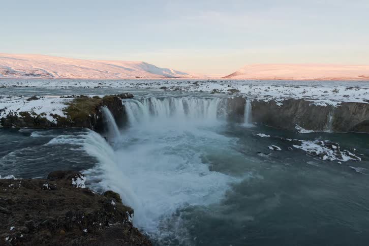 A wide crescentshaped waterfall cascading into a churning river set against a snowy mountainous landscapethe scene transitions from a soft pink dawn sky to a brighter sunlit morning as light gradually illuminates the icy cliffs and surrounding plai