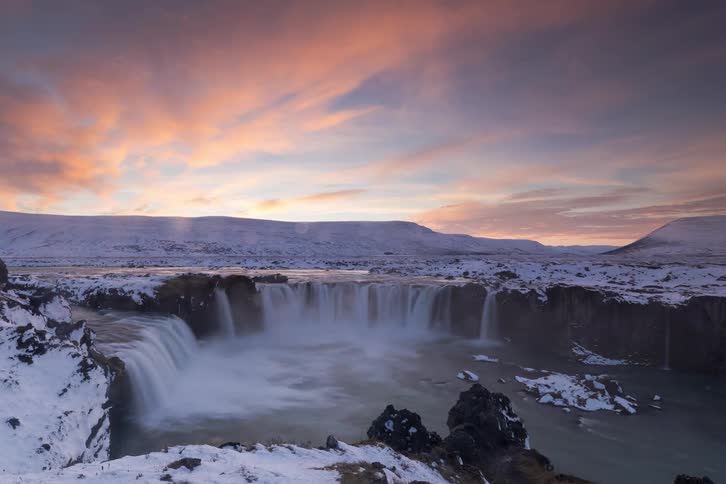 A wide winter landscape of a powerful waterfall flowing through a snowy canyon under a pink sunset sky