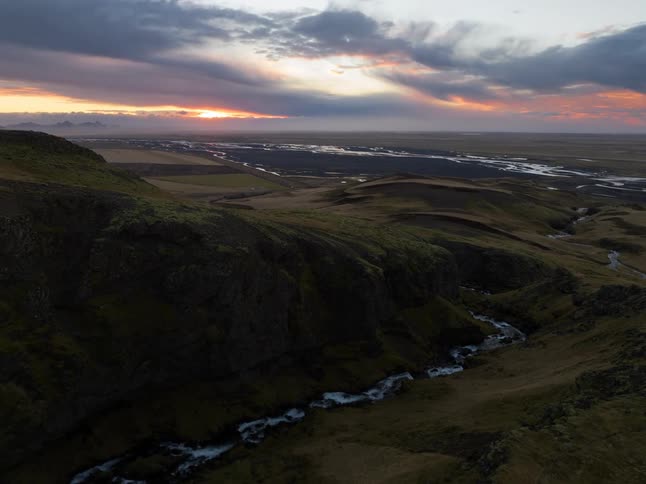Aerial wide of a dramatic waterfall cutting through rolling green hills and a winding river at sunset