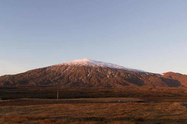 A mountain with a snowcovered peak in the badlands during fall with vast land covered in short brown grasses and some people camping from midday to sunset