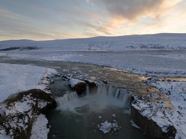 A high angle view shows a wide horseshoe waterfall flowing into a river surrounded by a vast snow covered plain under a pale sunset sky with distant white mountains on the horizon