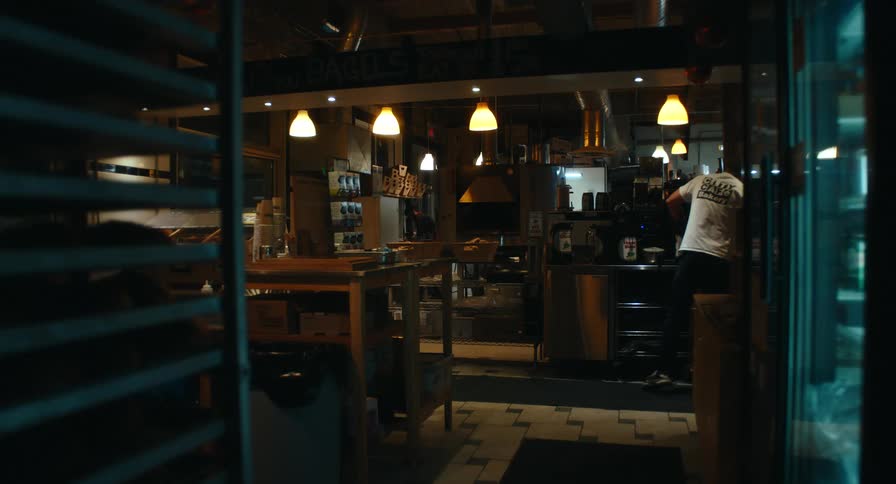 A view from a doorway shows a baker in a white tshirt working in a professional kitchen with warm pendant lights stainless steel equipment and shelves filled with supplies during early morning hours