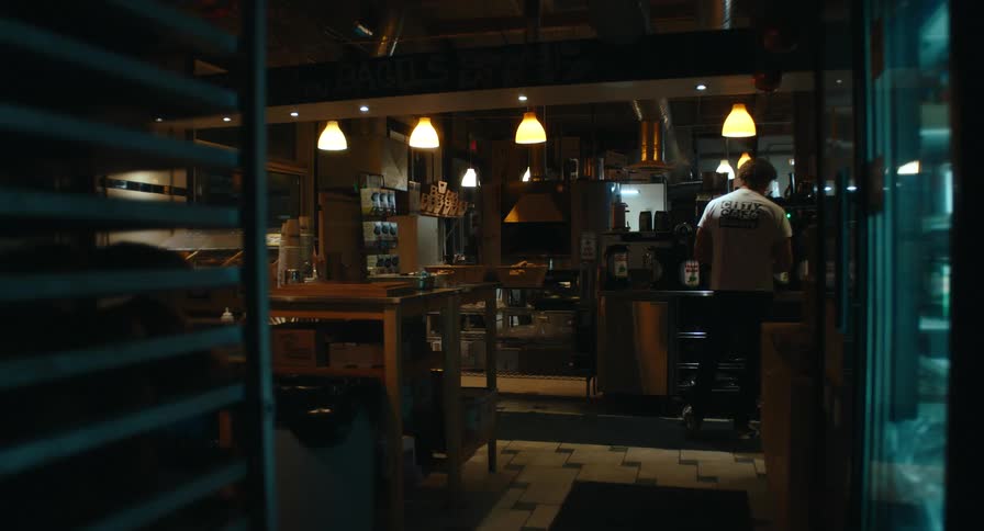A worker tidies a dimly lit cafe kitchen at night framed through shelves under warm industrial lighting