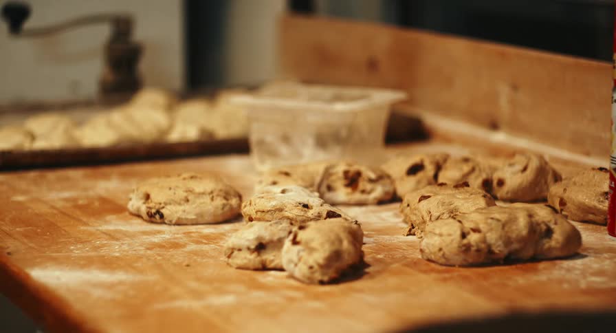 A close up view shows several portions of raw dough mixed with raisins or fruit sitting on a flourdusted wooden workbench in a professional bakery