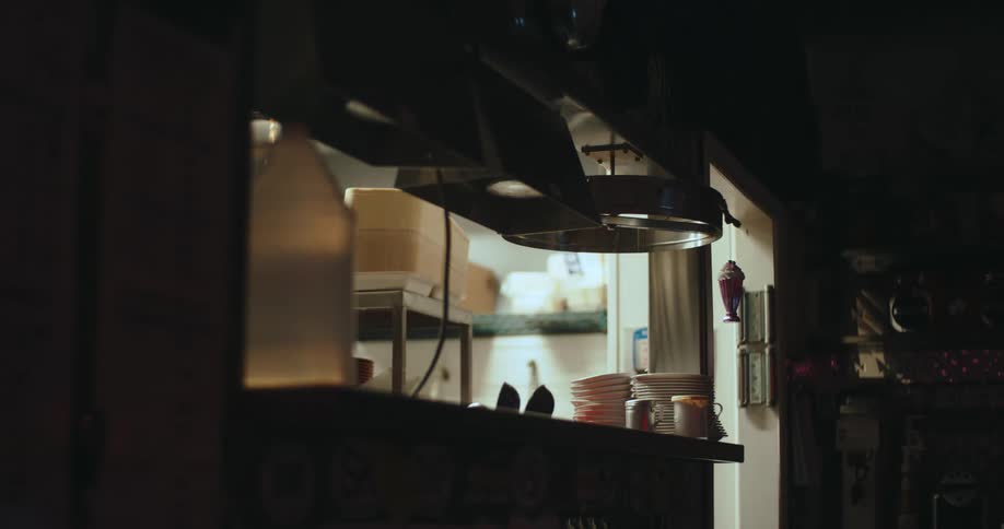 A low angle view looks through a dark diner service window where stacked white plates and a large plastic container sit on the counter under industrial heat lamps in a dimly lit commercial kitchen