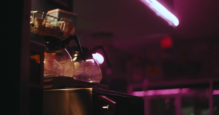 A close view shows glass coffee pots sitting on a brewing machine in a dimly lit cafe with a pink neon light glowing in the background