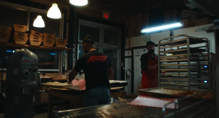 Two bakers work in a spacious commercial kitchen featuring hanging pendant lights and industrial equipment while preparing dough on wooden tables near large metal cooling racks and a heavy duty planetary mixer
