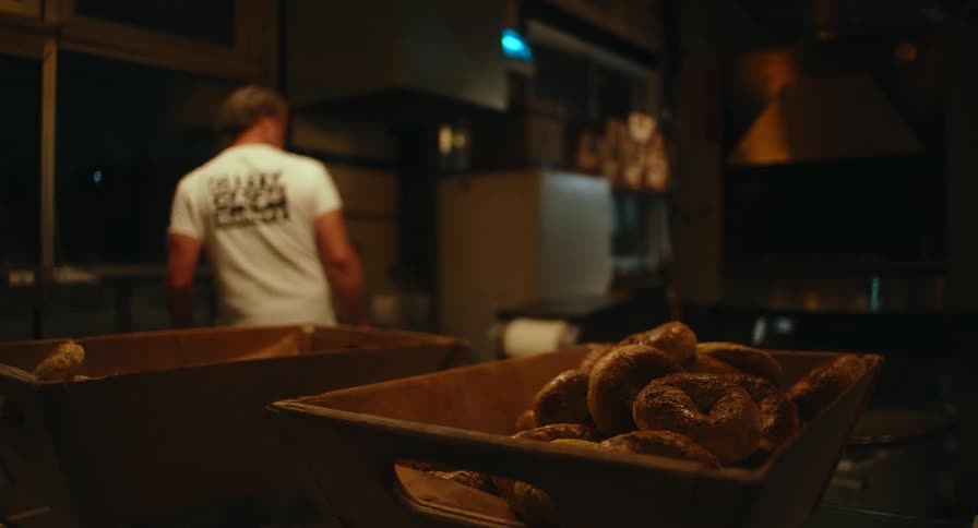 Freshly baked bread rests in wooden trays under warm golden light as a baker works quietly in the background of a cozy kitchen
