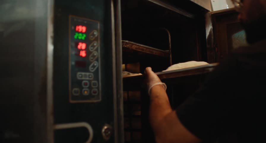 A man wearing a black shirt and white gloves slides a metal tray topped with raw dough into a large industrial oven featuring a glowing digital control panel on the side within a dark professional bakery