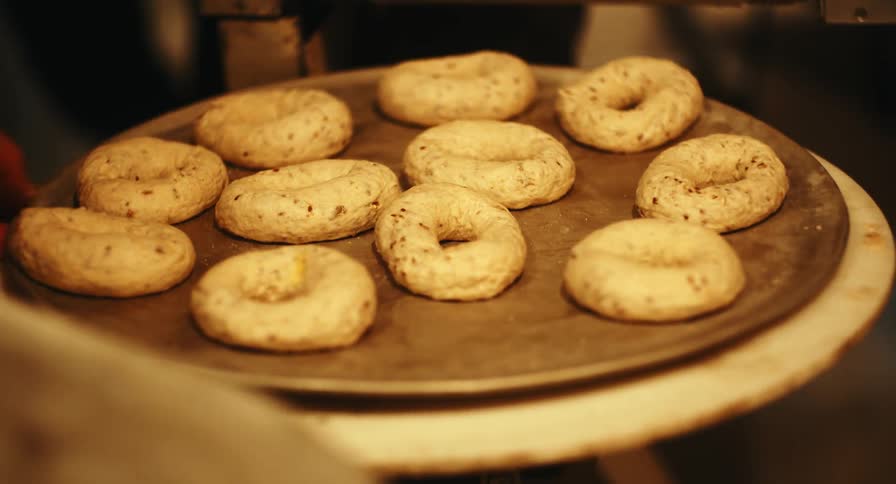 A bakers hand placing raw bagels onto a metal tray in a warm dimly lit kitchen