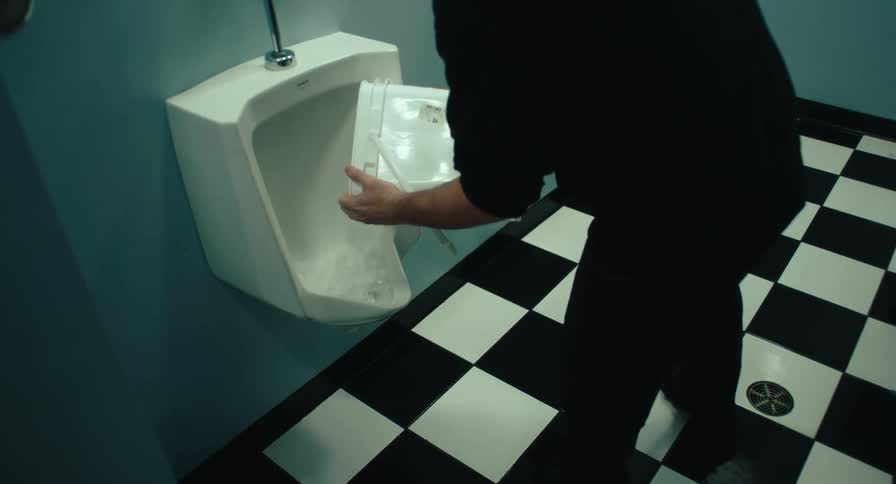 Janitor walks into a bathroom and pours a bucket of water into the urinal