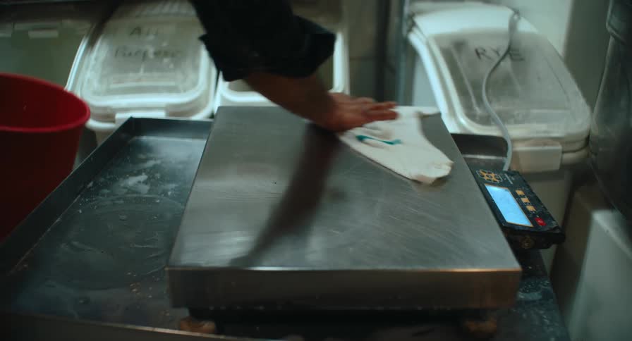 A close view shows a person in a dark long sleeve shirt using a white cloth to wipe down a large metal digital scale in a professional kitchen near a red bucket and bins of flour
