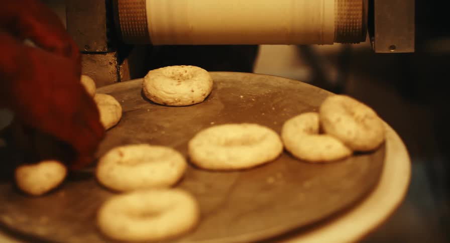 Raw bagels roll out of a machine and bakers handle them