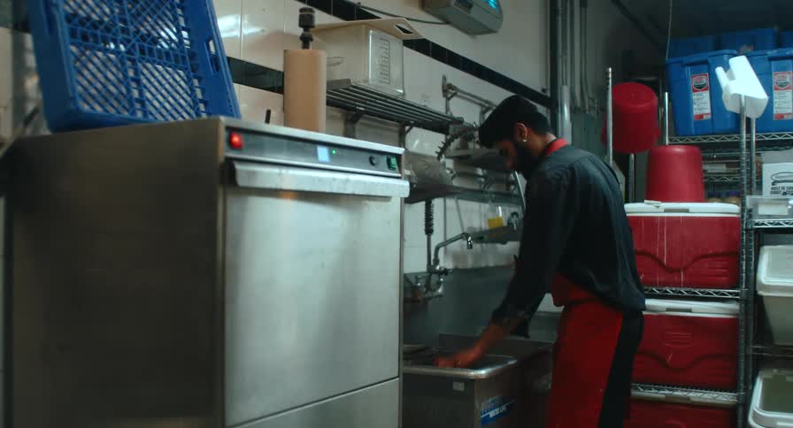 A kitchen worker in a red apron washes dishes at a commercial sink