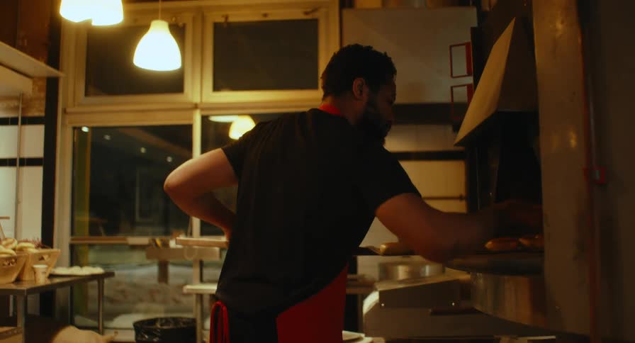 A baker reaches into a glowing oven under warm kitchen lights while another man works in the background