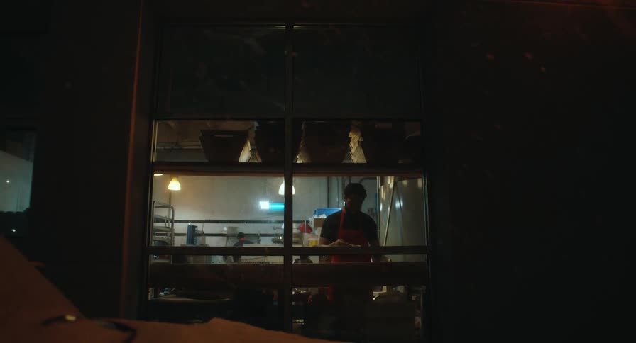A view through a window shows two bakers working in a professional kitchen with warm overhead lighting during the early morning hours
