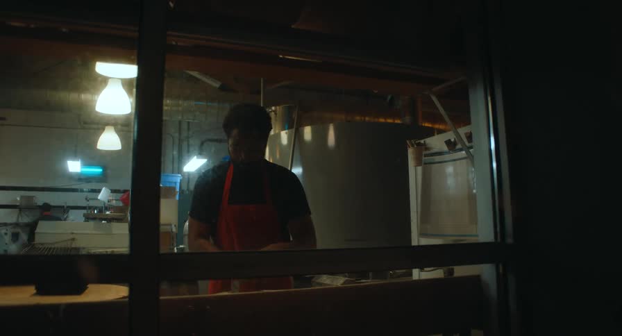 A view through a window shows a baker wearing a black shirt and red apron working at a wooden counter in a professional kitchen with warm overhead lighting during early morning hours