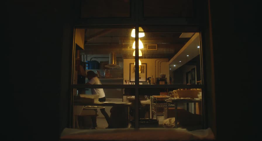 A view through a window shows a baker in a white shirt working in a professional kitchen with warm lights and stainless steel equipment during the early morning hours