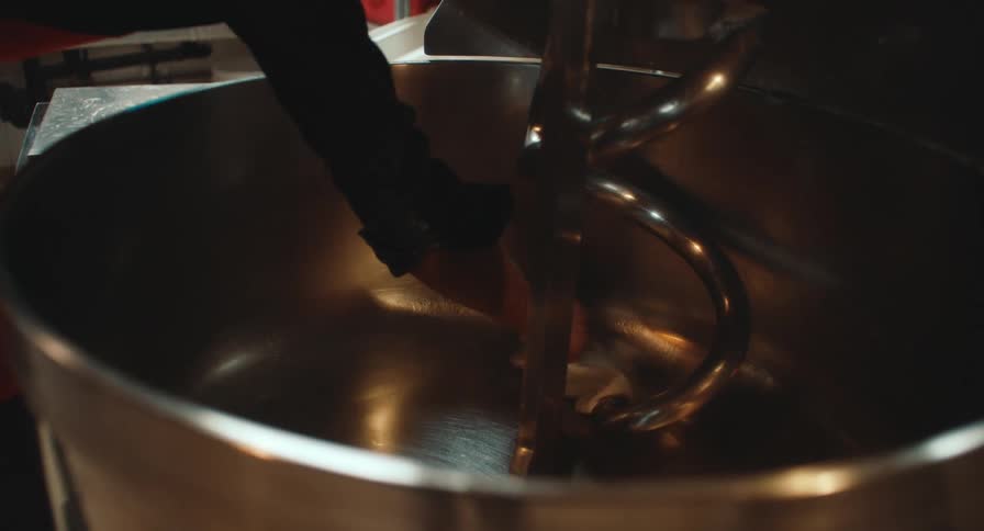 A professional baker in a black shirt and red apron cleans industrial equipment including a large planetary mixer and a professional oven with a glowing digital display inside a commercial kitchen workspace