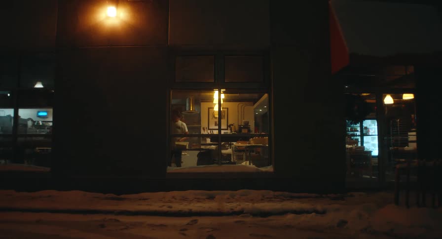 A baker works inside a warmly lit bakery at night seen through a window with snow on the ground outside