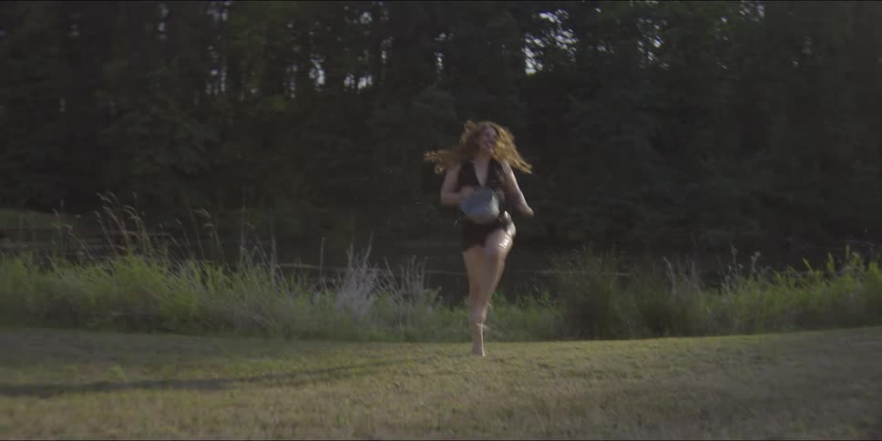 A shirtless man in black shorts runs across a grassy field while a woman in a dark dress chases him and throws a bucket of water creating a large splash against the backdrop of a dense forest