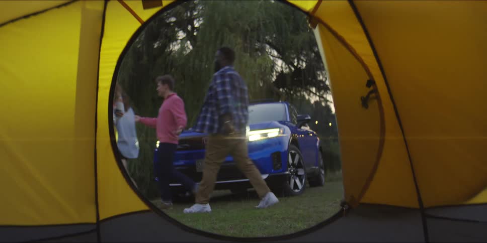 A group of friends walks past a blue suv at a campsite framed by the circular opening of a yellow tent