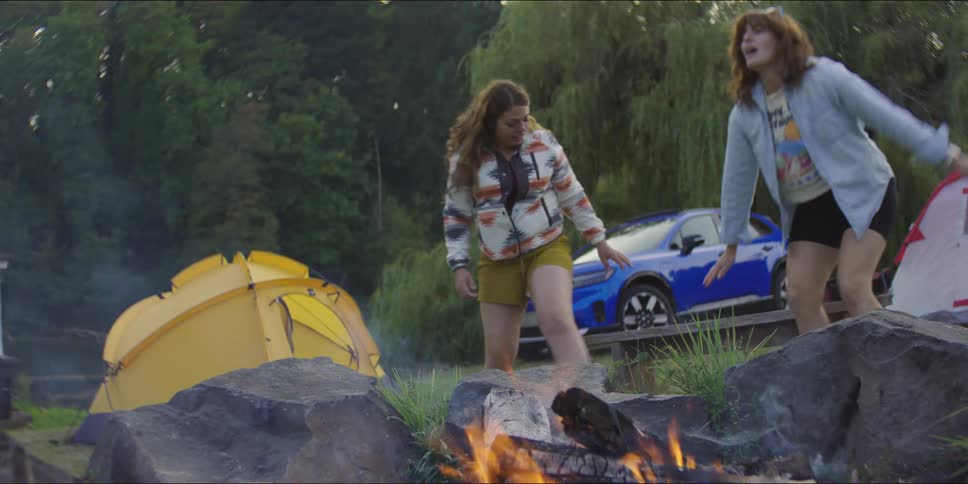 Two women set up a wooden bench and sit down to talk by a crackling campfire at a campsite with a blue suv and yellow tent in the background