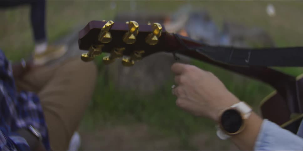 A person wearing a white watch and a ring plays an acoustic guitar while sitting near a blurry campfire in a grassy field during the evening