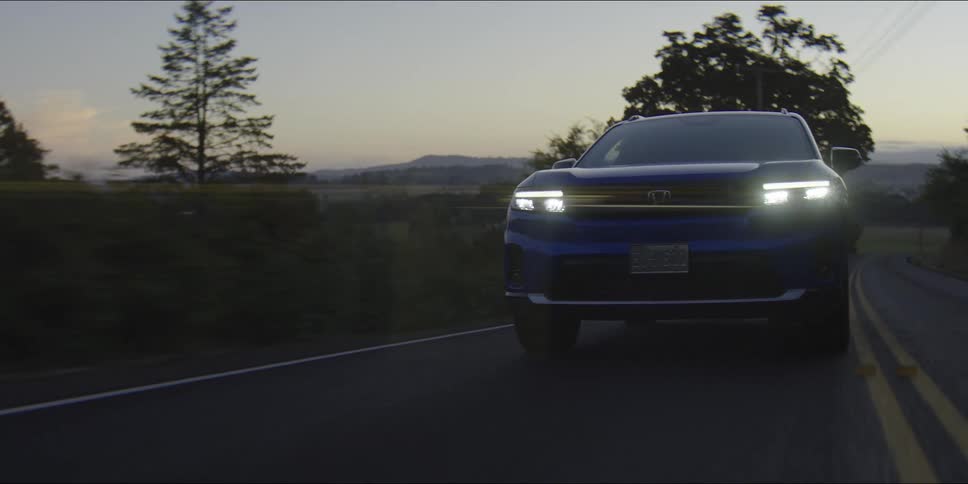 Blue personal car driving on a road surrounded by sparse trees with mountains and hills on the background outdoors during dusk or dawn