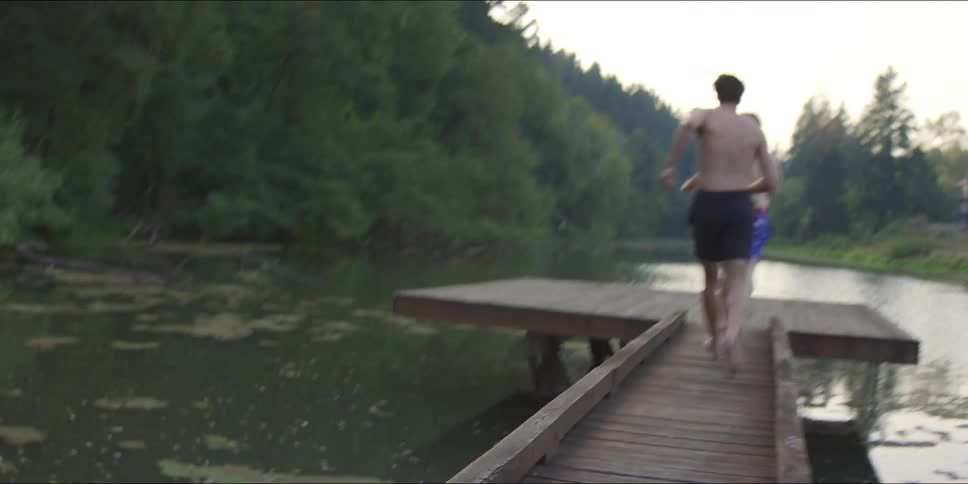 Two men and a woman run across a wooden pier and prepare to jump into a quiet lake surrounded by dense green forest and a house in the distance during a bright summer afternoon