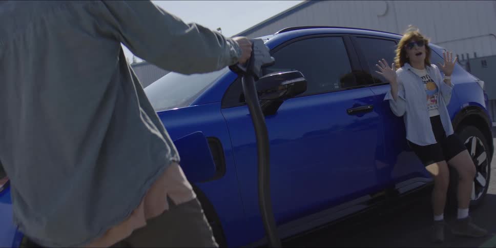 Young adult woman wearing sunglasses standing against a blue car and young adult man removing the gas pump and pointing at her like a gun to make her laugh outdoors during day time