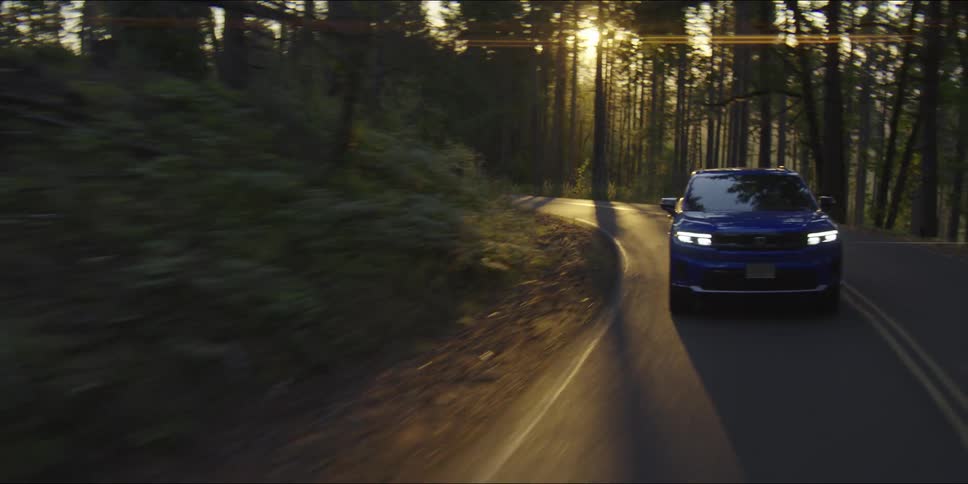 A blue suv drives along a winding road through a dense forest as the sun shines through the trees during sunset