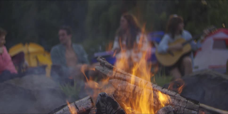 Flames crackle in the foreground of a campfire as friends gather in soft focus behind it sharing musicstrummng a guitar and conversation beneath the evening sky