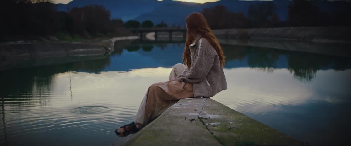 Young adult redhead woman sitting next to a pond throwing pebbles at the water with trees and mountains on the background outdoors during dusk or dawn