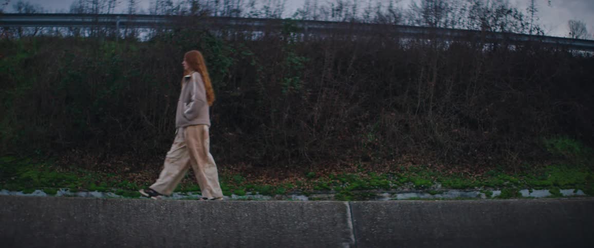 A side view shows a woman with long wavy red hair wearing a beige fleece jacket and tan pants walking along a concrete path next to a line of dense bushes and a highway during twilight