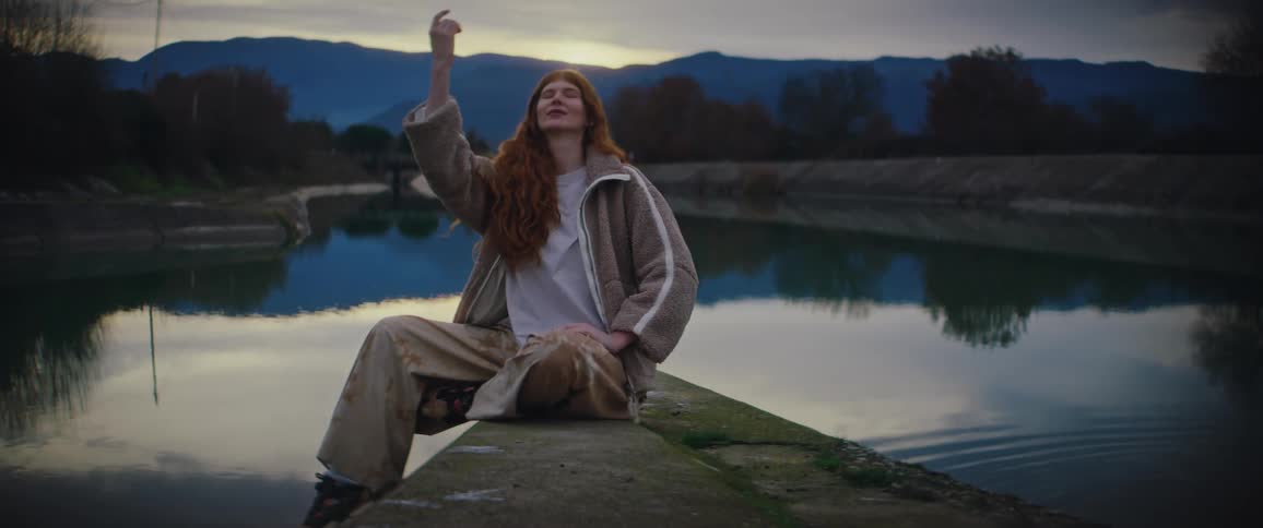 A woman with long red hair sits on a concrete ledge by a calm river and throws a small stone into the water while the surrounding mountains and evening sky reflect on the surface during a quiet twilight in the countryside