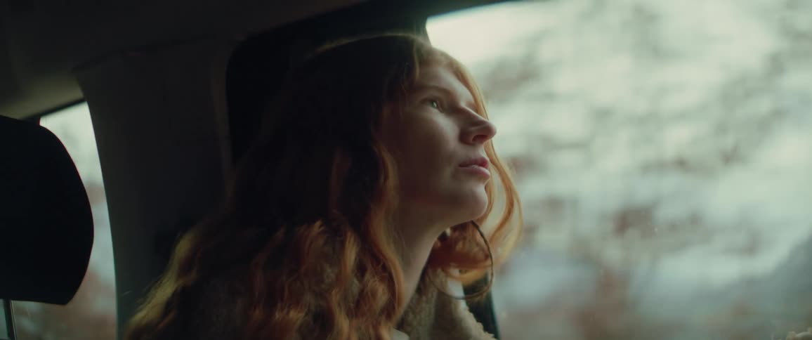 A teenage girl with long red hair sits inside a car looking out the window at a blurry forest landscape filled with bare trees and distant mountains as soft light hits her face