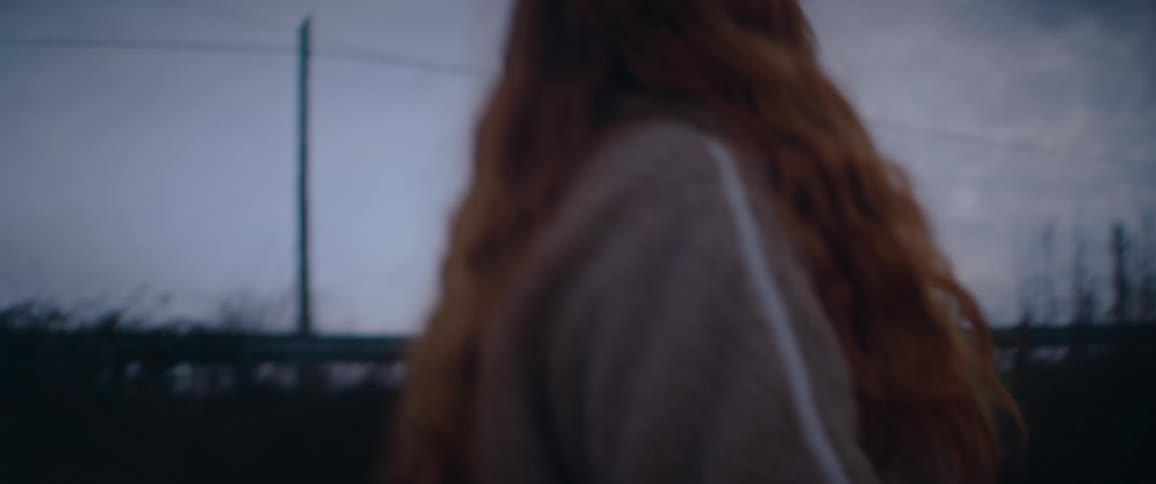 A redhaired woman stands beneath a moody overcast sky her gaze turned to the distance as wind as she touches her hair