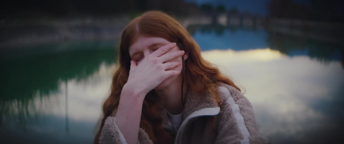 Young adult redhead woman sitting and touching her face with a sad facial expression in front of a pond with mountains and trees on the background outdoors during dusk or dawn