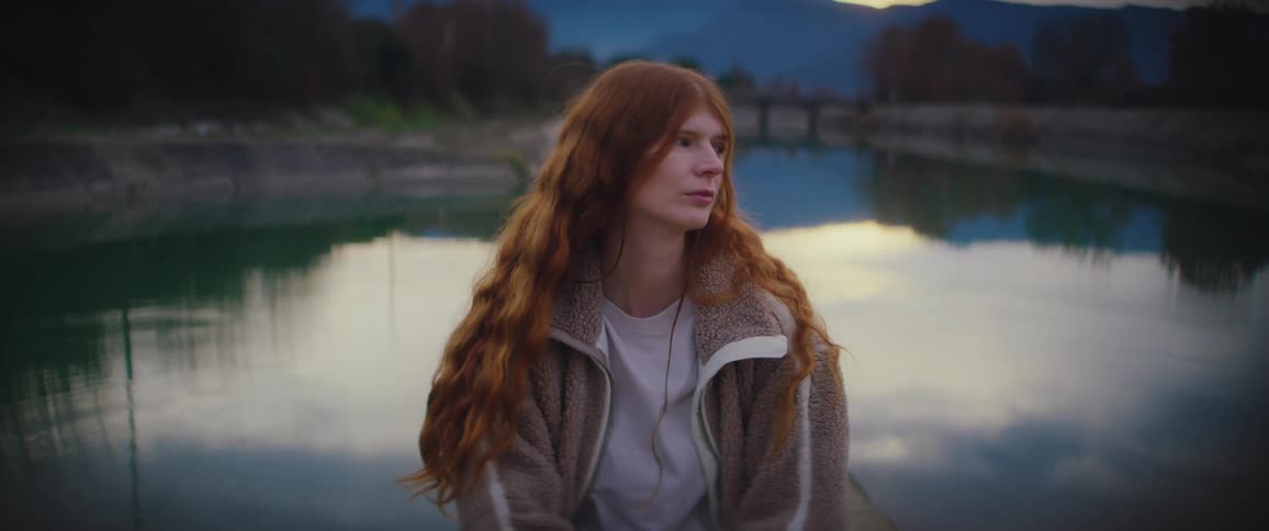 A woman with long ginger hair wearing a fleece jacket sits by a still river at dusk looking at her phone and then away toward the horizon while mountains and a bridge are visible in the soft evening light