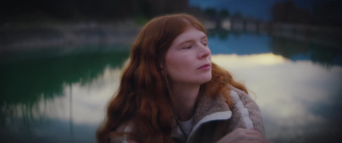 A woman with long red hair looks around while sitting near a lake