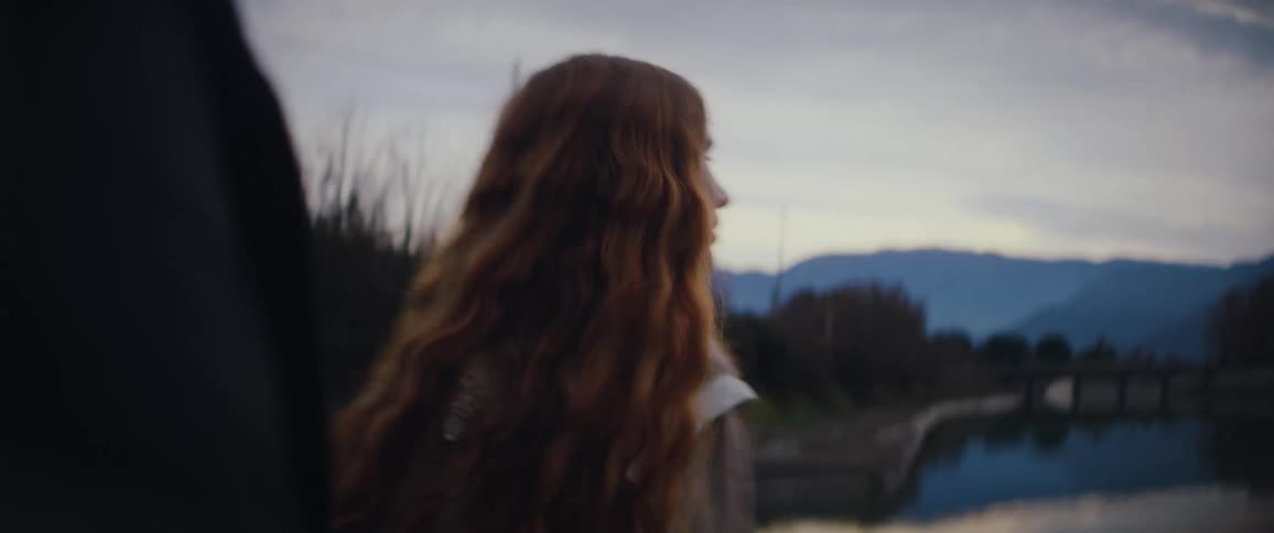 A young woman with long wavy red hair is skipping stones across a calm body of water during twilight the scene captures her leaning over to toss a stone and then standing up to watch its ripples spread across the surface
