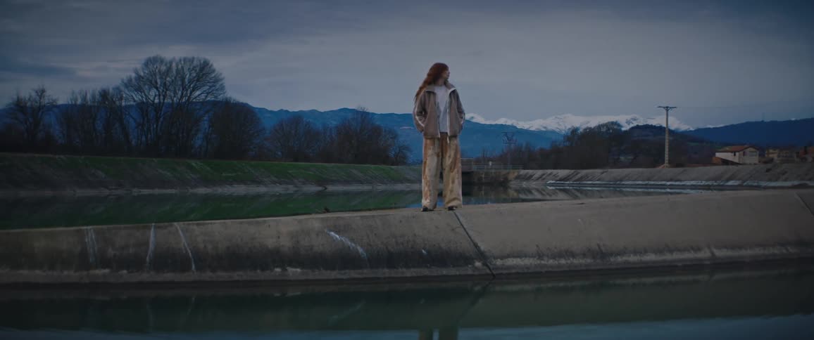 A young woman in beige stands on a concrete divide between two canals with snow capped mountains in the distance