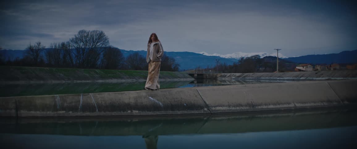 A woman in a beige jacket and patterned pants walks along a concrete ledge above a reflective pool of water with snow capped mountains and a dark cloudy sky in the background